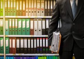 Businessman holding a briefcase standing by shelves of organized binders.